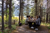 A group of participants in the forest overlooking a lake, sitting at a picnic table eating lunch.